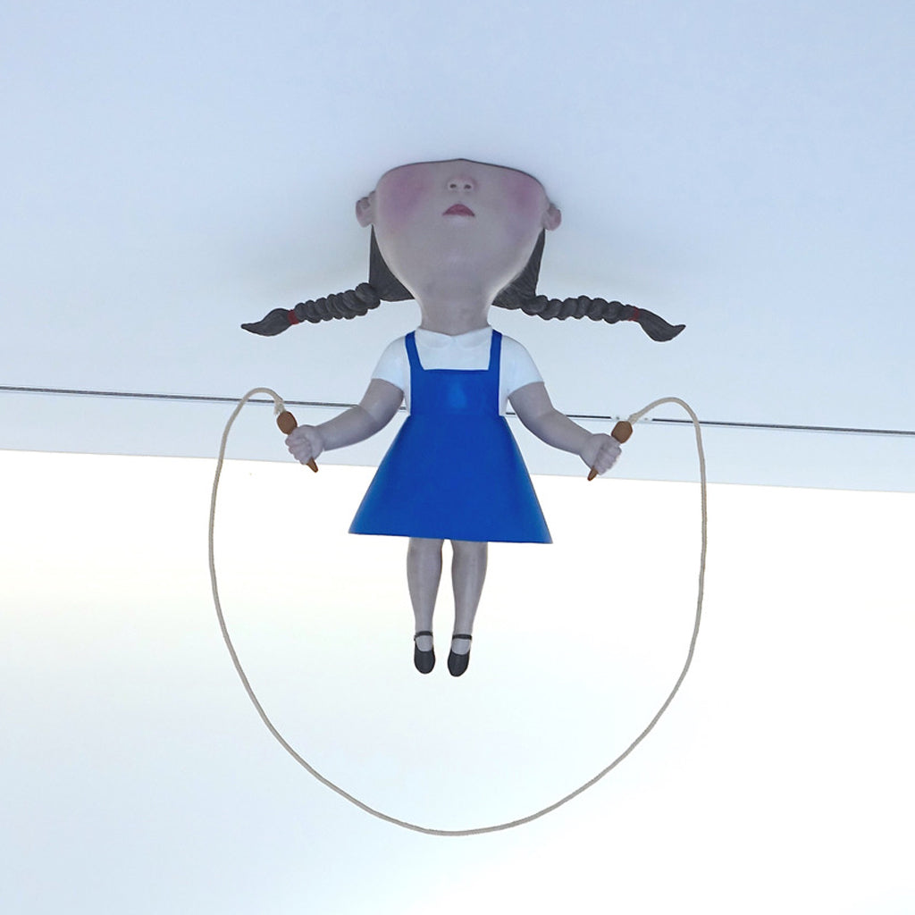 A sculpture of a girl in a blue pinafore with braids jumping rope, mounted upside down on a ceiling