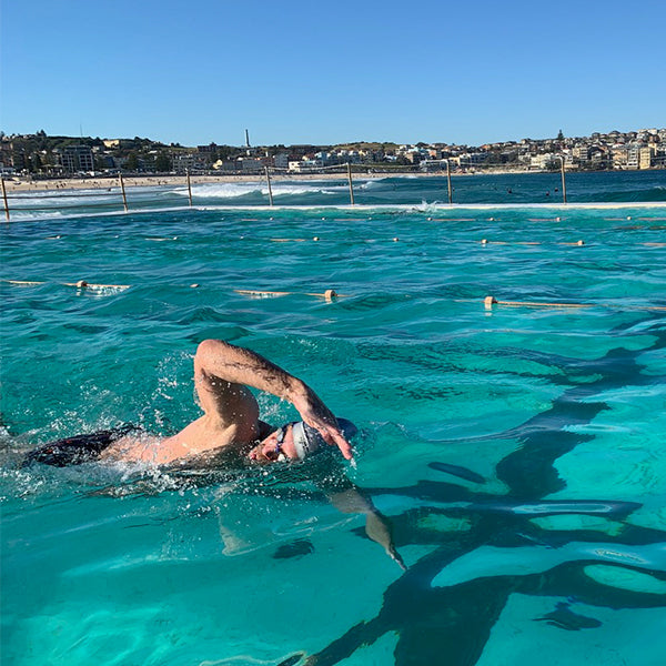 A person in a swim cap and goggles swims freestyle in a turquoise ocean pool beside a coastal town