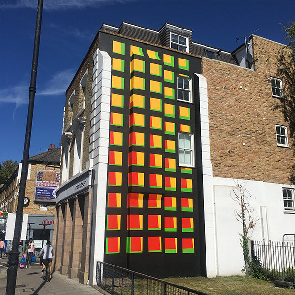 A black building wall painted with a grid of red, orange and yellow squares with green edges, beside a brick building