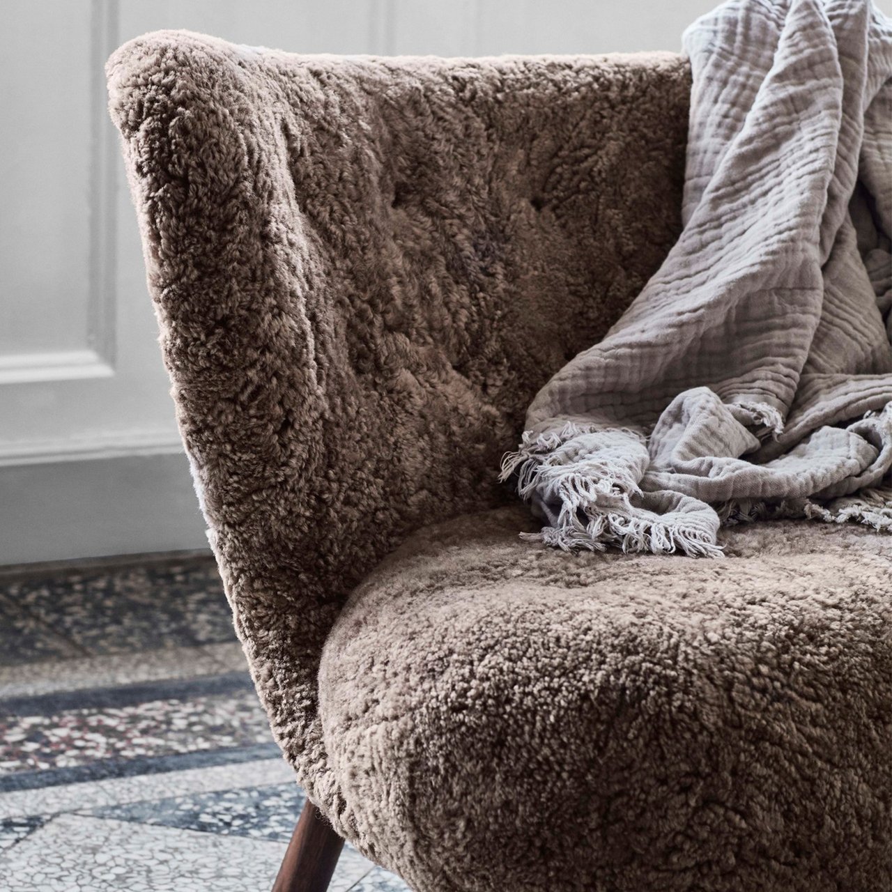 Close-up of a brown shearling armchair with a pale grey throw draped over its arm, on a stone tile floor