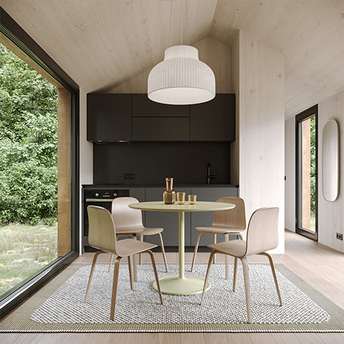 A round pale green table with four light wood chairs under a ribbed white pendant in a compact wood-lined kitchen