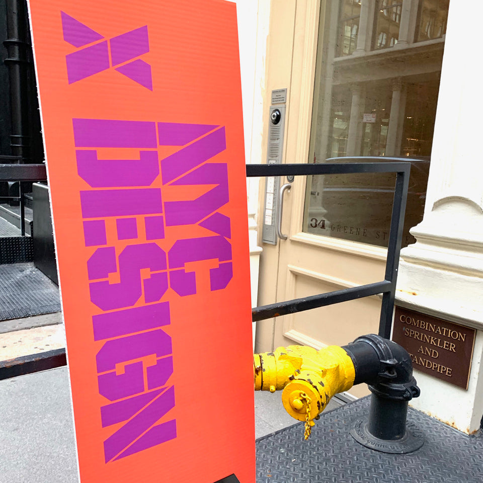 A tall orange sign with purple block letters reading NYC Design stands beside a yellow standpipe outside a cream doorway
