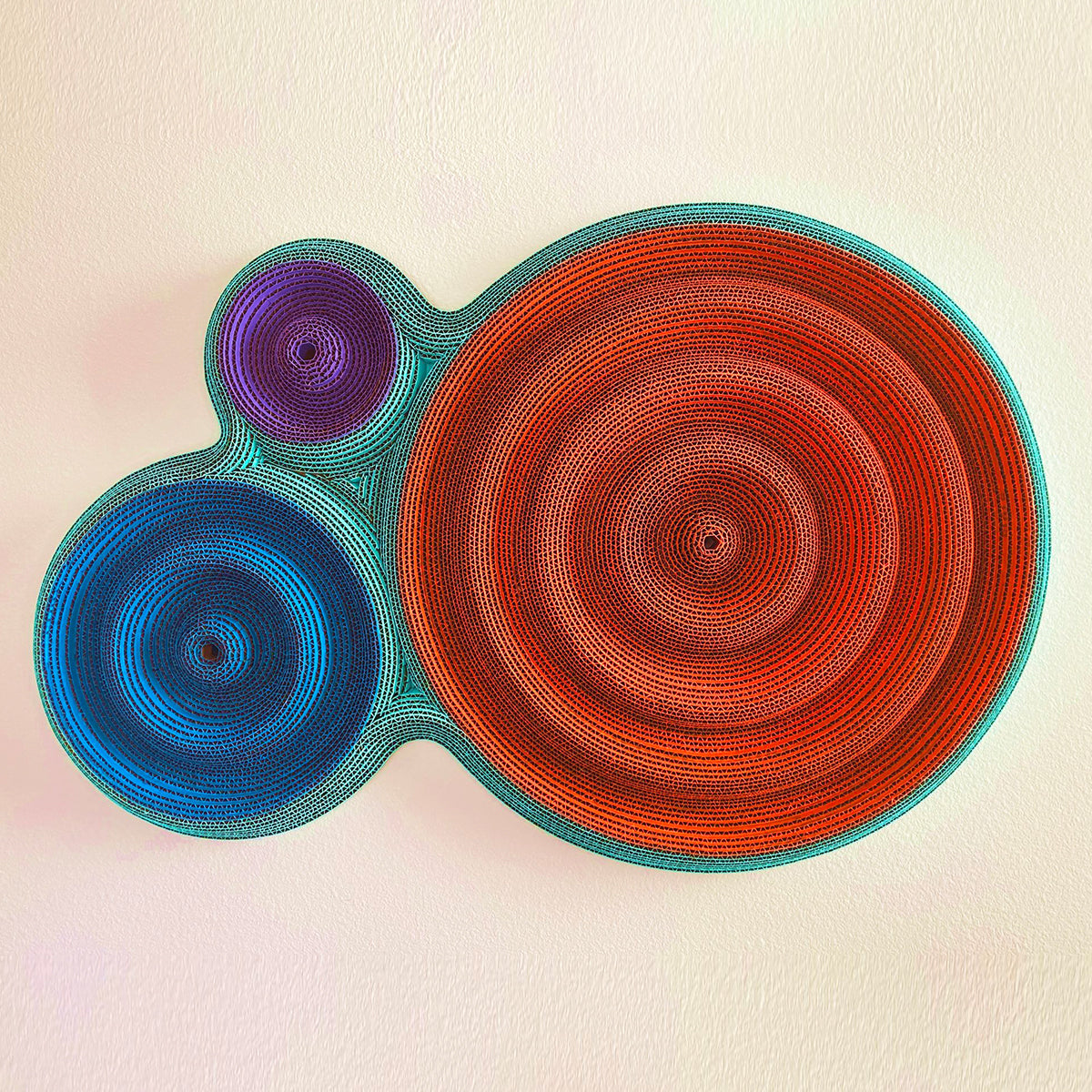 A wall-mounted sculpture of three joined circles in orange, blue and purple, with concentric ridged rings and teal edges