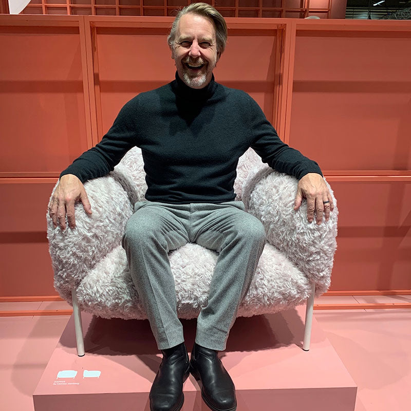 A shaggy pale grey armchair with slim white legs, with a person seated in it on a pink display platform