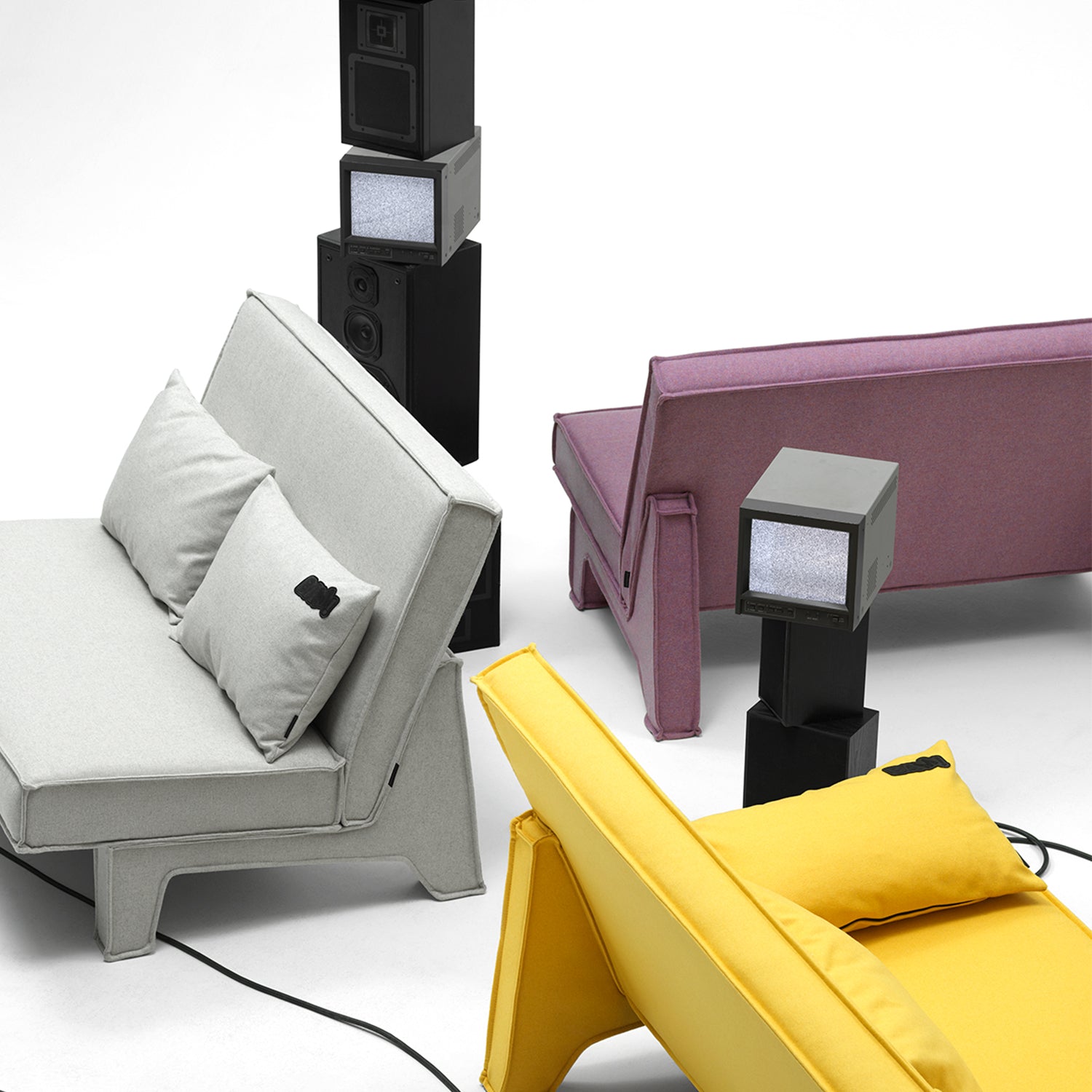 Three low-backed sofas in pale grey, mauve and yellow fabric among stacked speakers and small TVs