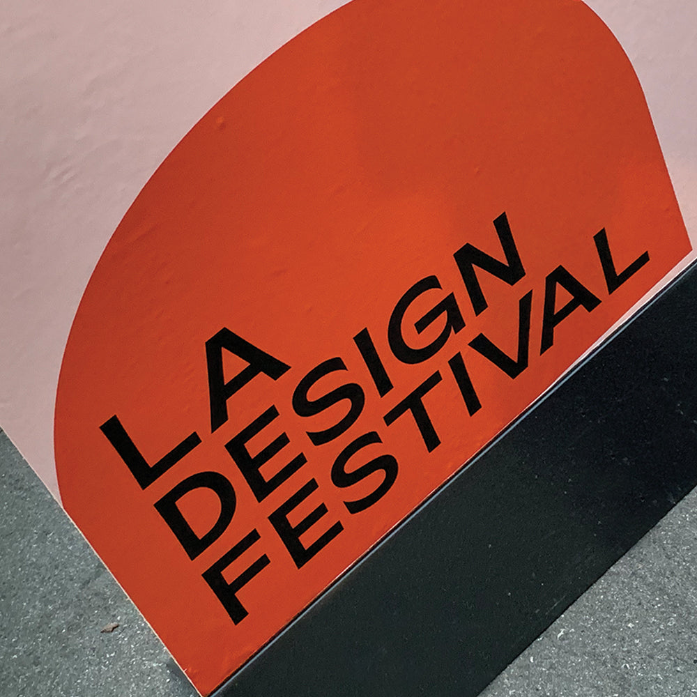 Angled sign with black text reading LA Design Festival on an orange arch against a pale pink wall