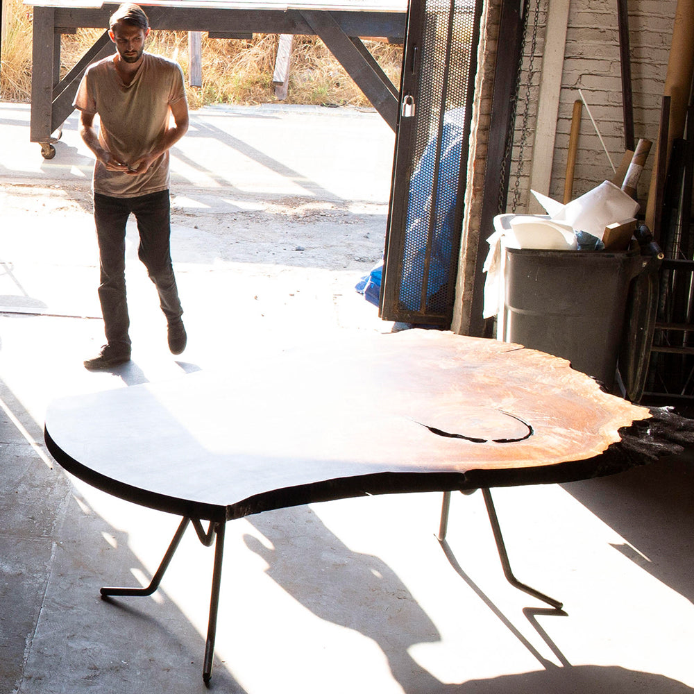 A large live-edge wood slab table on thin black metal legs in a workshop doorway, with a person walking toward it