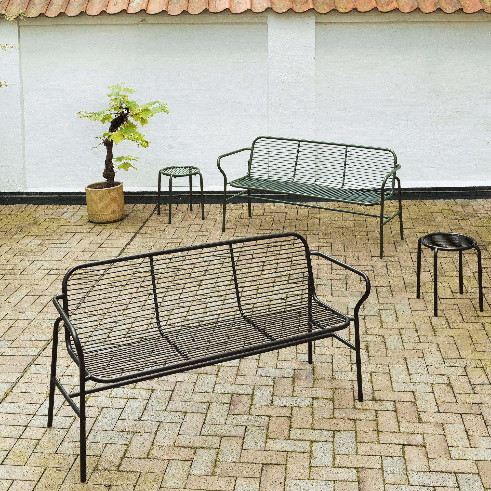 Black metal mesh Vig dining bench with slender frame on light brick patio.
