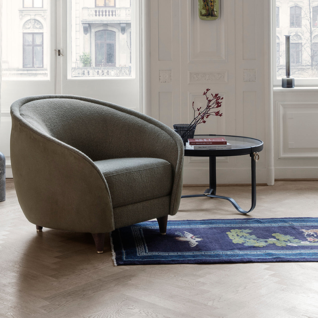 A round glass-topped coffee table on a dark curved metal base beside a green armchair in a bright room