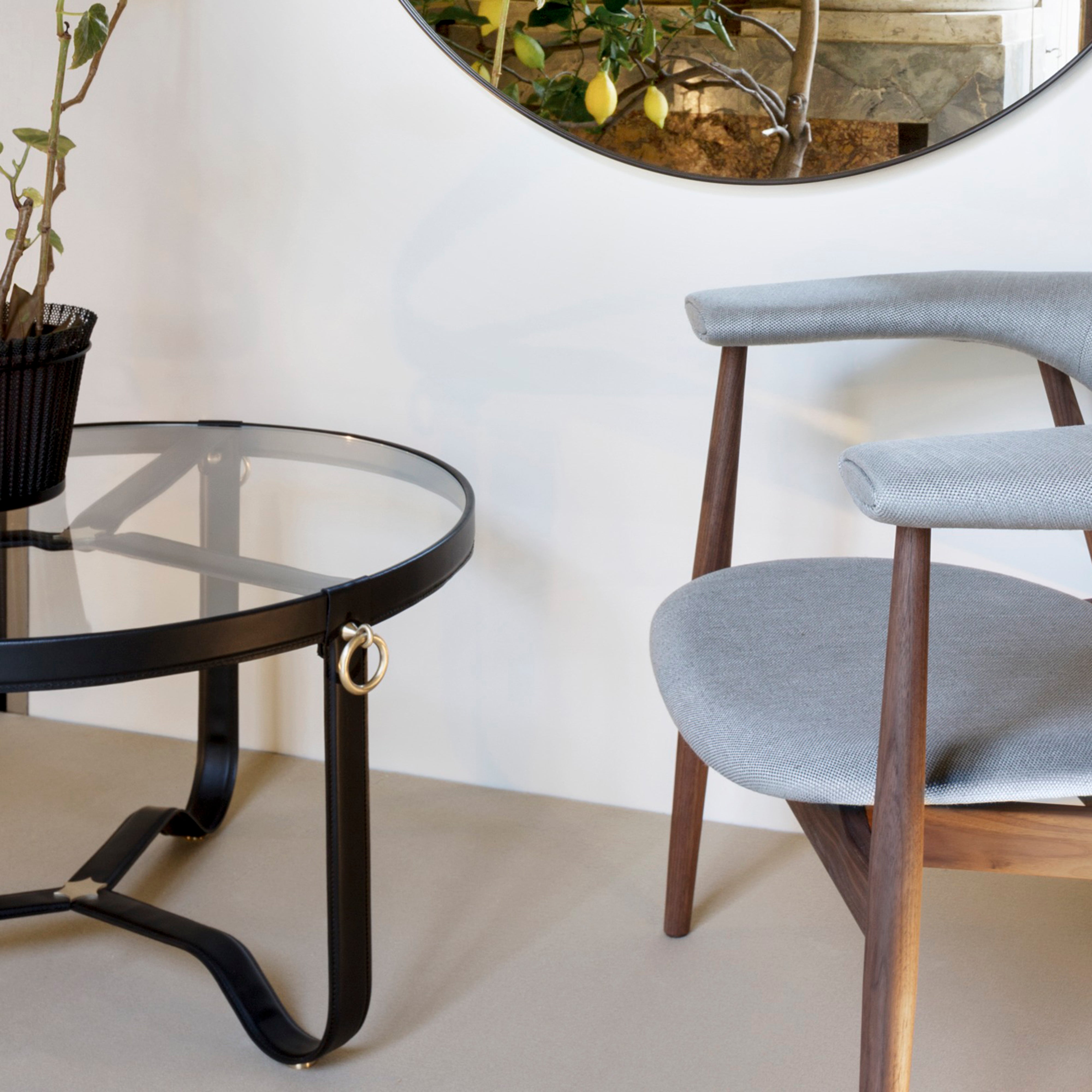A round glass coffee table with black stitched leather straps and a brass ring, beside a grey chair