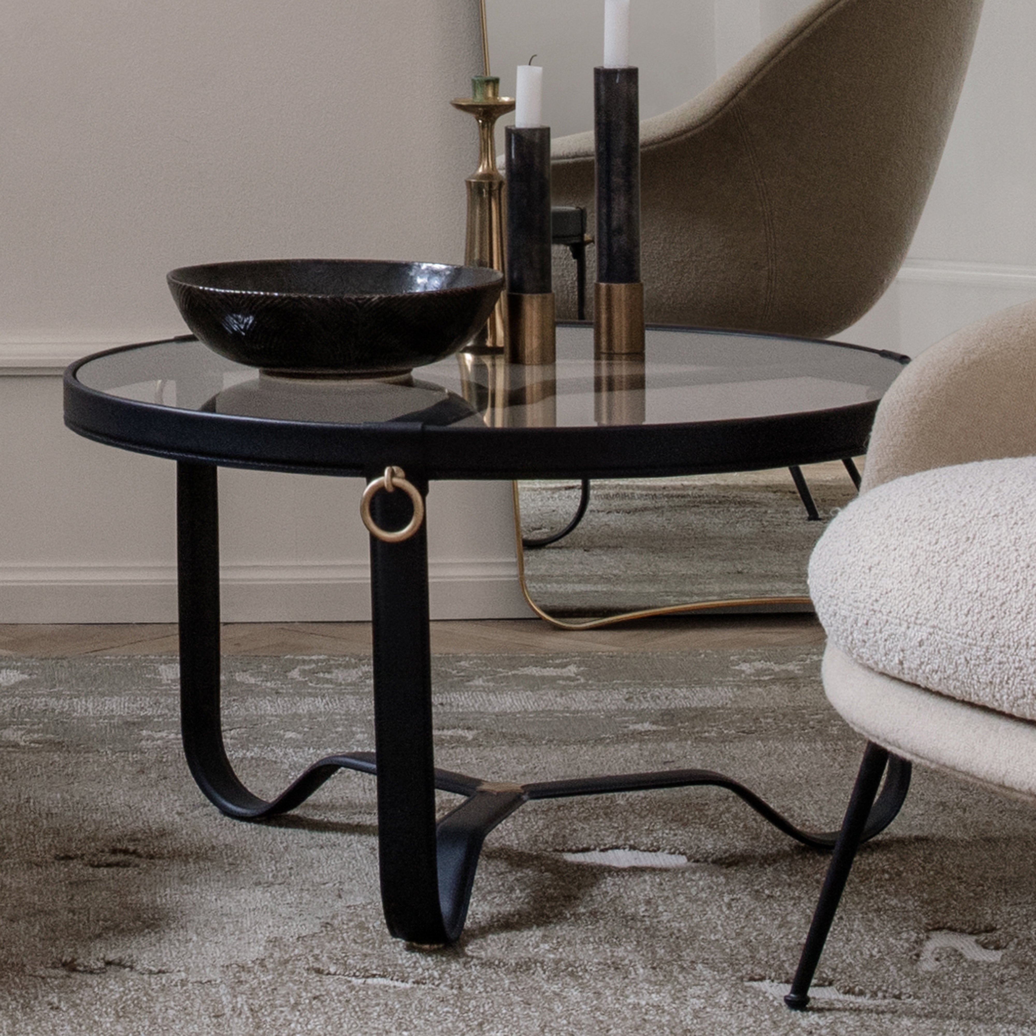 A round glass coffee table with black leather-wrapped legs and a brass ring, beside cream armchairs
