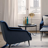 A round glass-topped table with a black frame, brass table lamp and glass bottles, beside a navy armchair