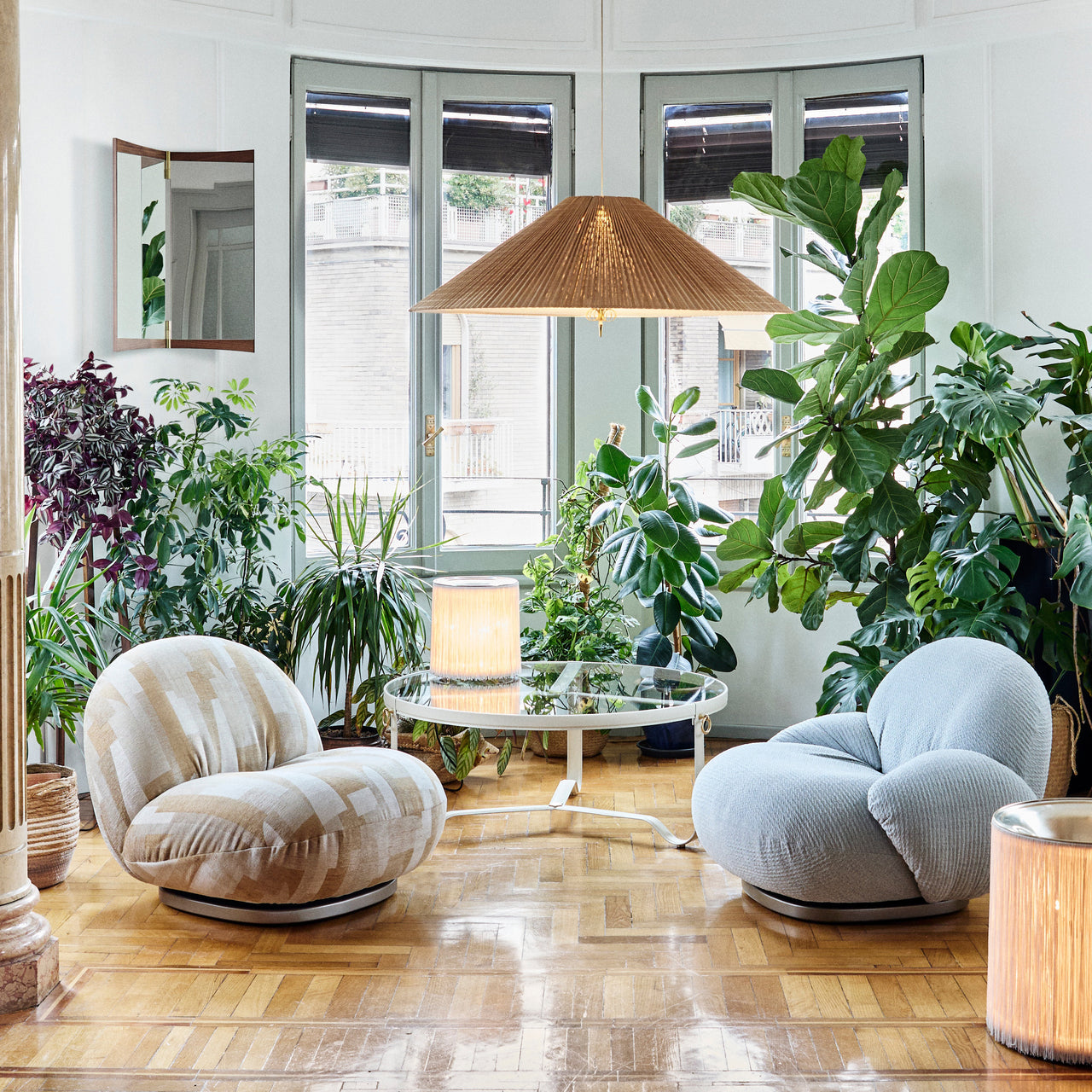 A round glass coffee table with a thin white frame between two rounded armchairs among large houseplants