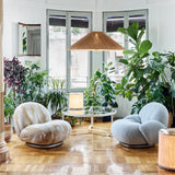 A round glass coffee table with a thin white frame between two rounded armchairs among large houseplants