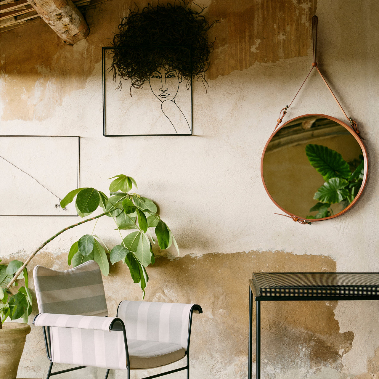 A round mirror with a tan leather strap hangs on a weathered plaster wall beside a striped chair