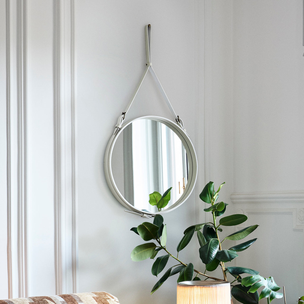 A round wall mirror with a pale leather strap and buckles, hung on a white panelled wall beside a green plant