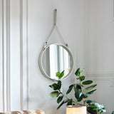 A round wall mirror with a pale leather strap and buckles, hung on a white panelled wall beside a green plant