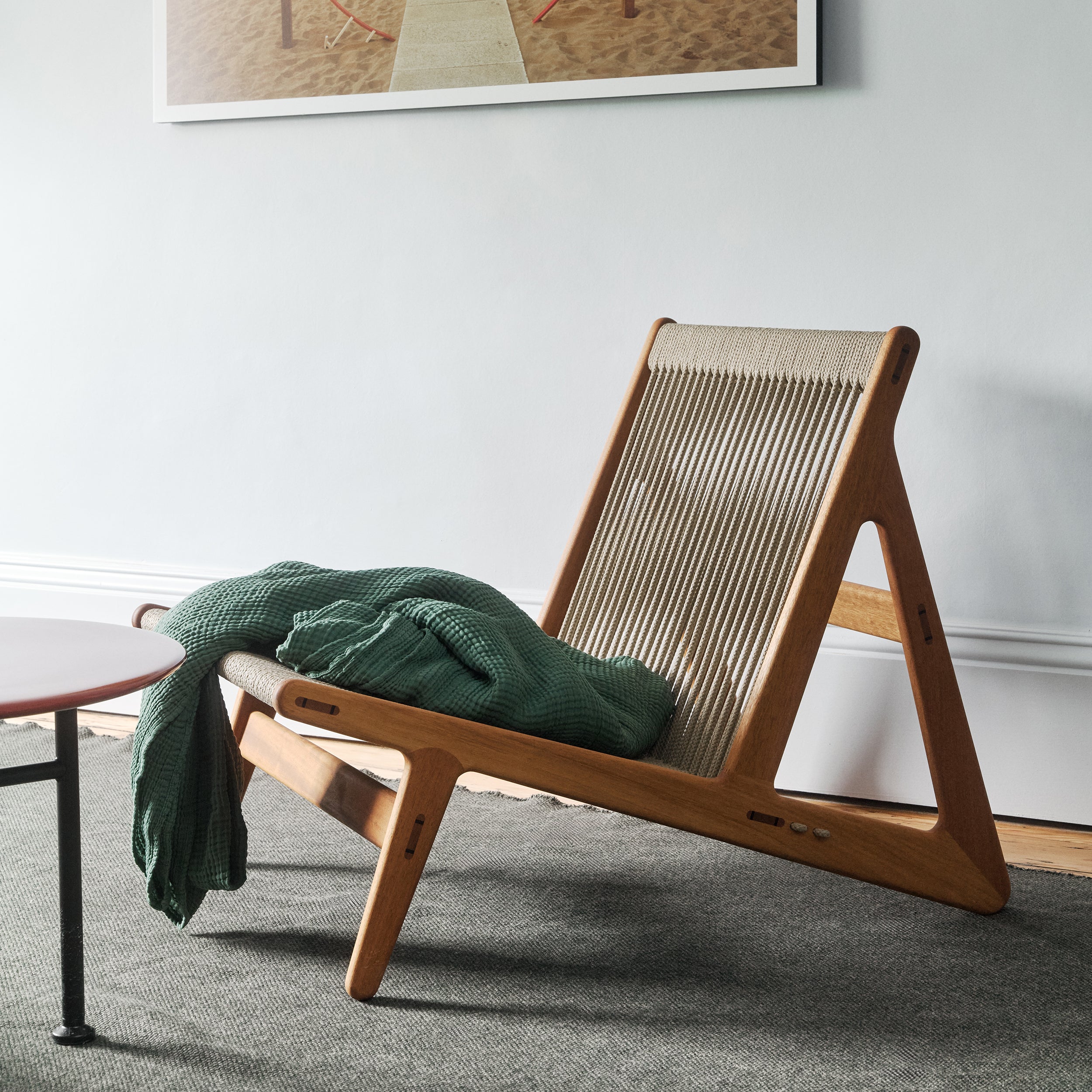 A low lounge chair with a light wood frame and woven rope back, a green throw over the seat