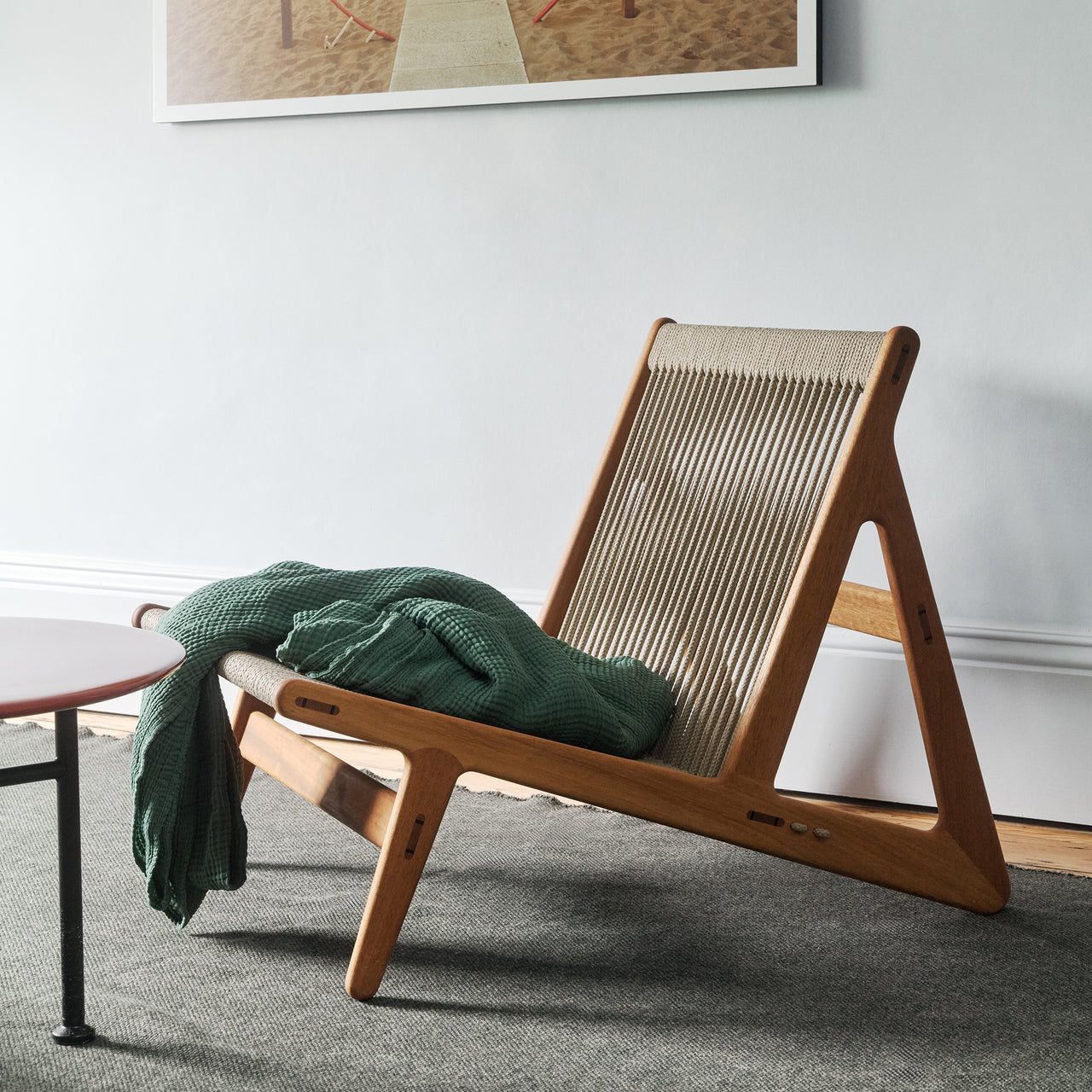 A low lounge chair with a light wood frame and woven rope back, a green throw over the seat