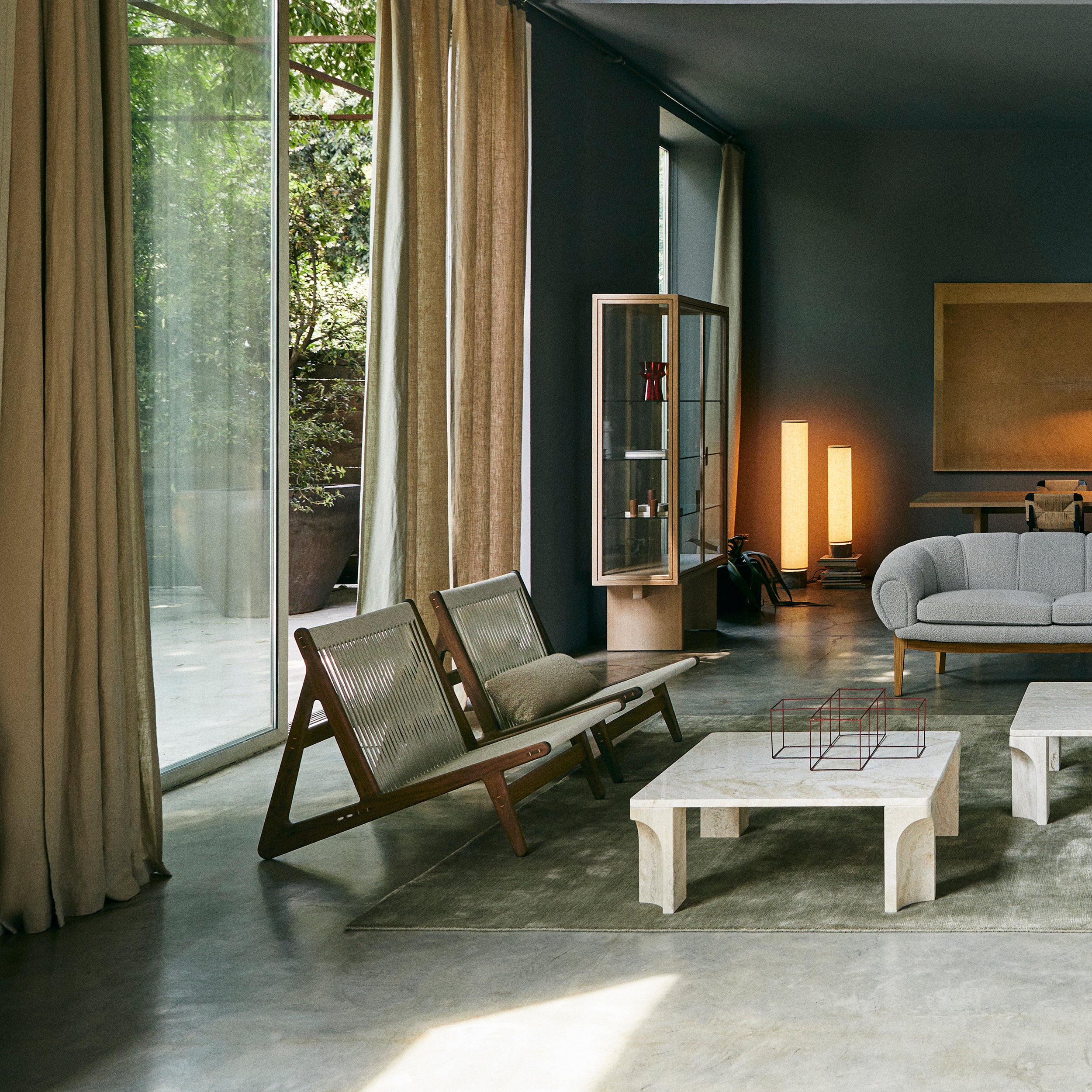 Two low wooden lounge chairs with woven cord backs in a concrete-floored room with linen curtains