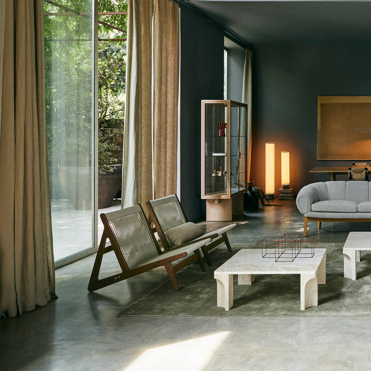 Two low wooden lounge chairs with woven cord backs in a concrete-floored room with linen curtains