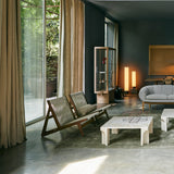 Two low wooden lounge chairs with woven cord backs in a concrete-floored room with linen curtains
