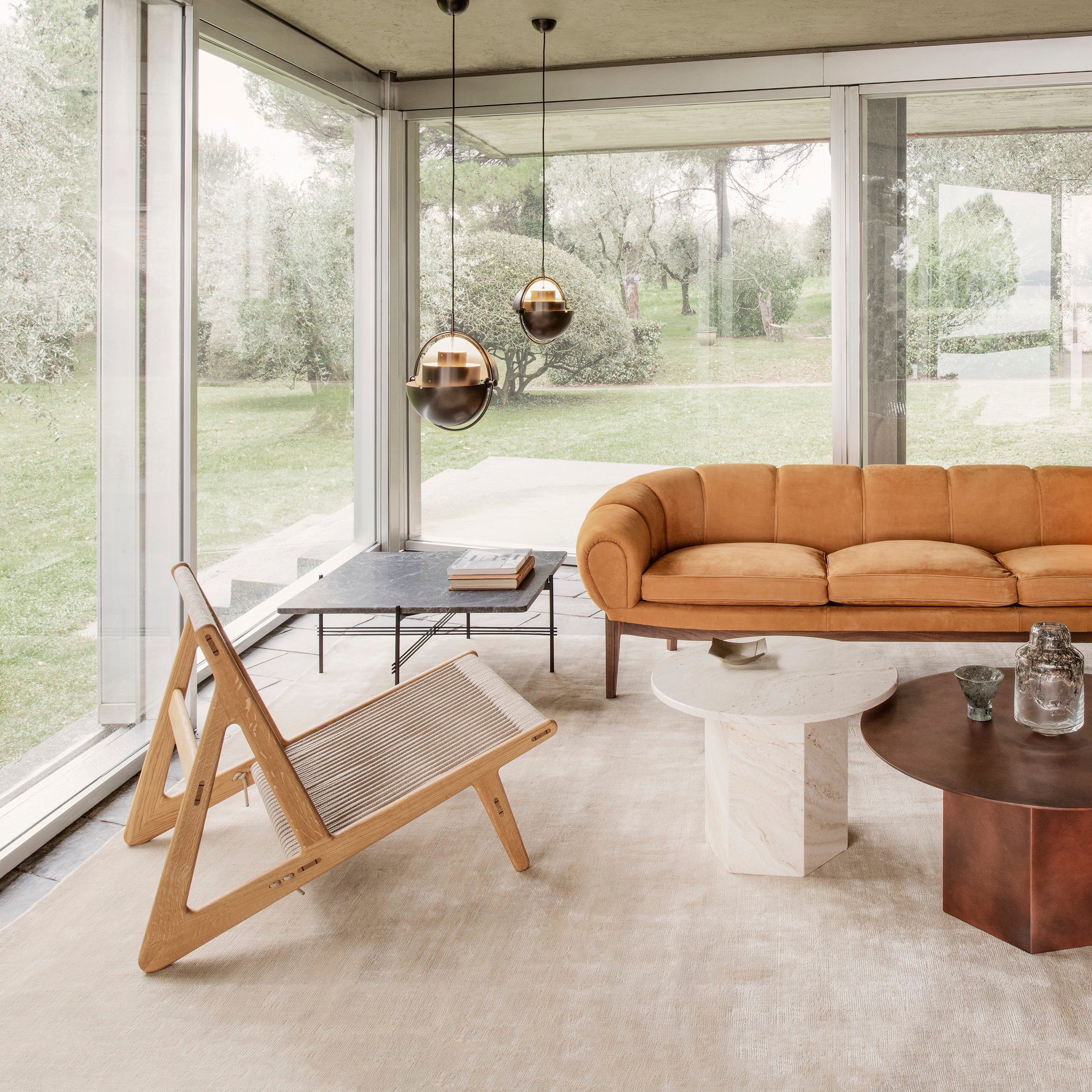 A light wood lounge chair with a rope seat in a glass-walled room with a tan sofa and round tables