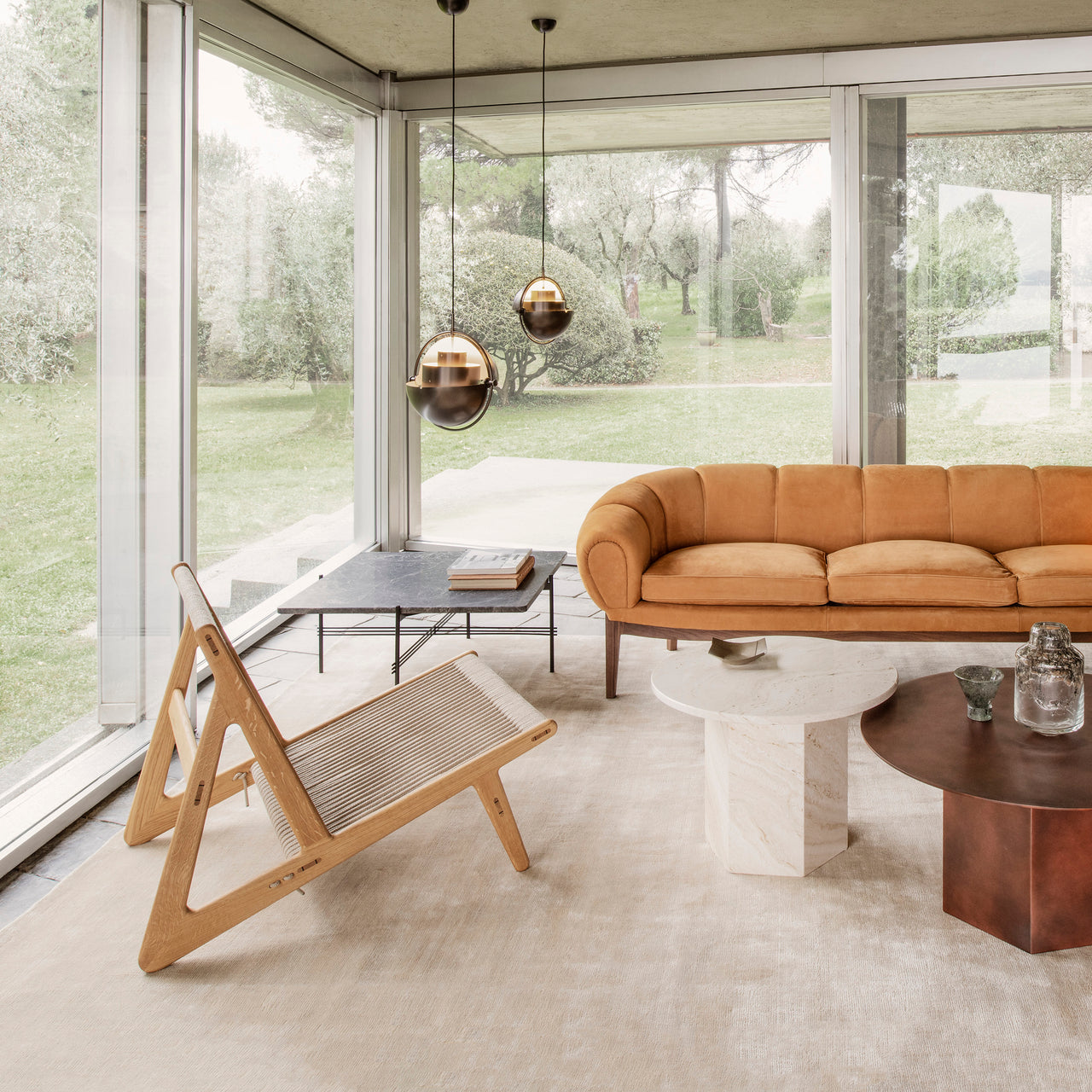A light wood lounge chair with a rope seat in a glass-walled room with a tan sofa and round tables