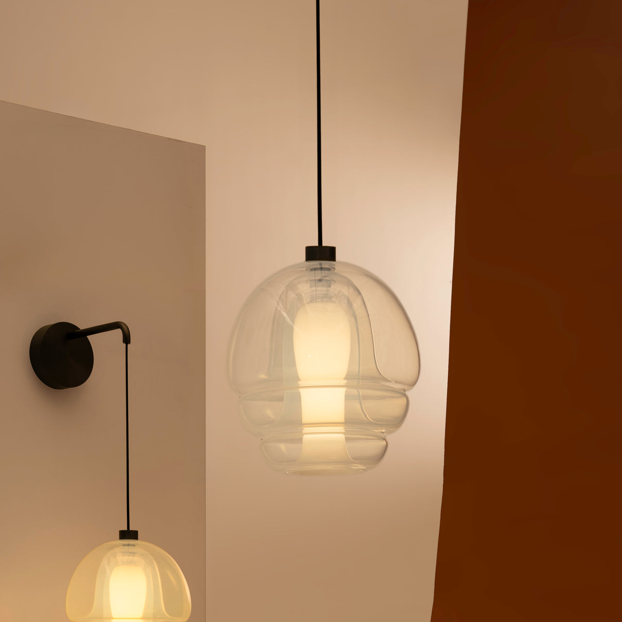 A clear glass pendant with a glowing white inner core hangs on a black cord, with a wall-mounted version nearby