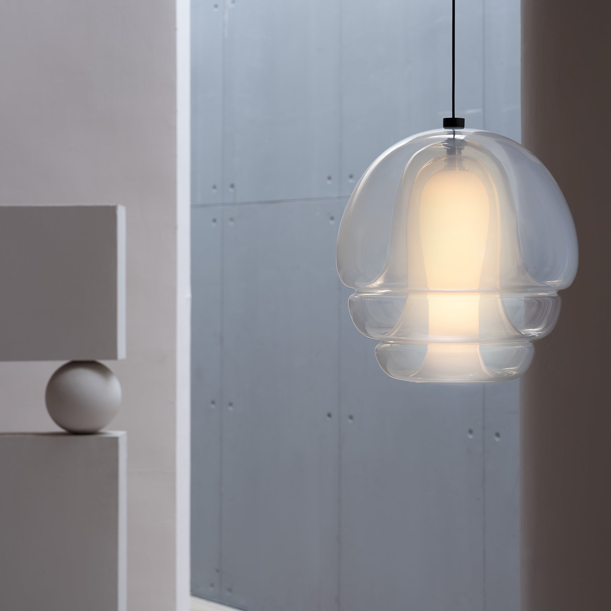 A glowing glass pendant with clear layered tiers around a frosted core, hanging against a grey concrete wall