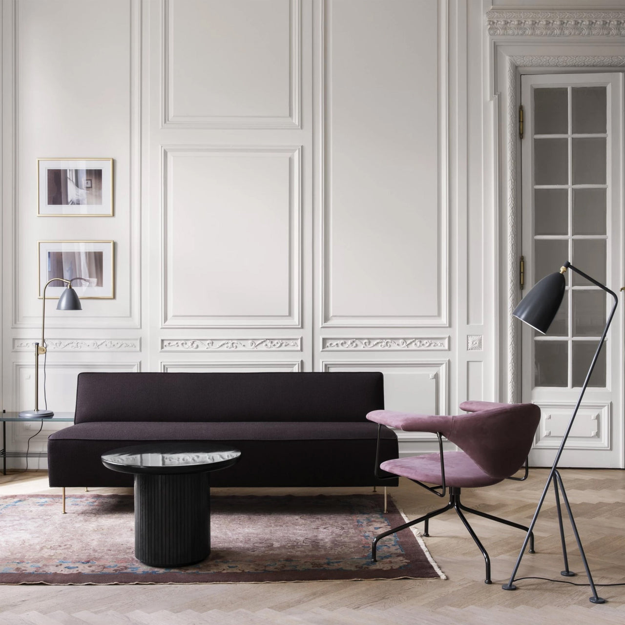 A low dark aubergine sofa on slim brass legs in a room with white panelled walls and a pink armchair