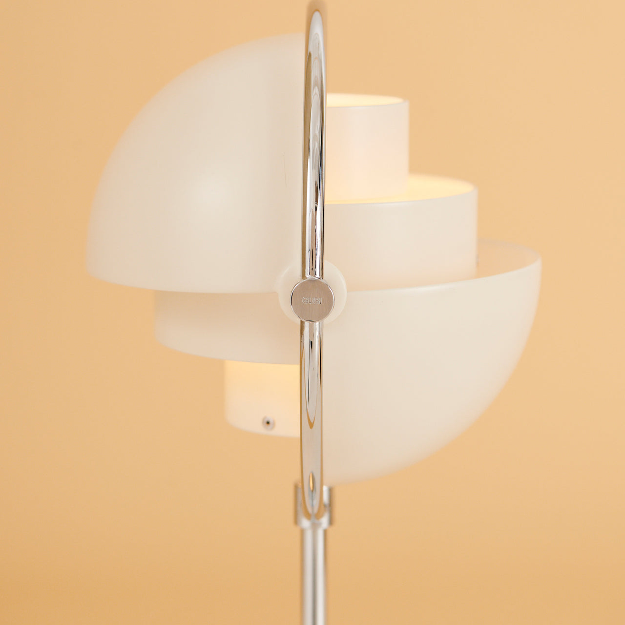 Close-up of a white lamp with stacked half-dome shades pivoting on a polished chrome arc, against a peach wall