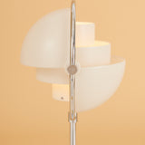 Close-up of a white lamp with stacked half-dome shades pivoting on a polished chrome arc, against a peach wall