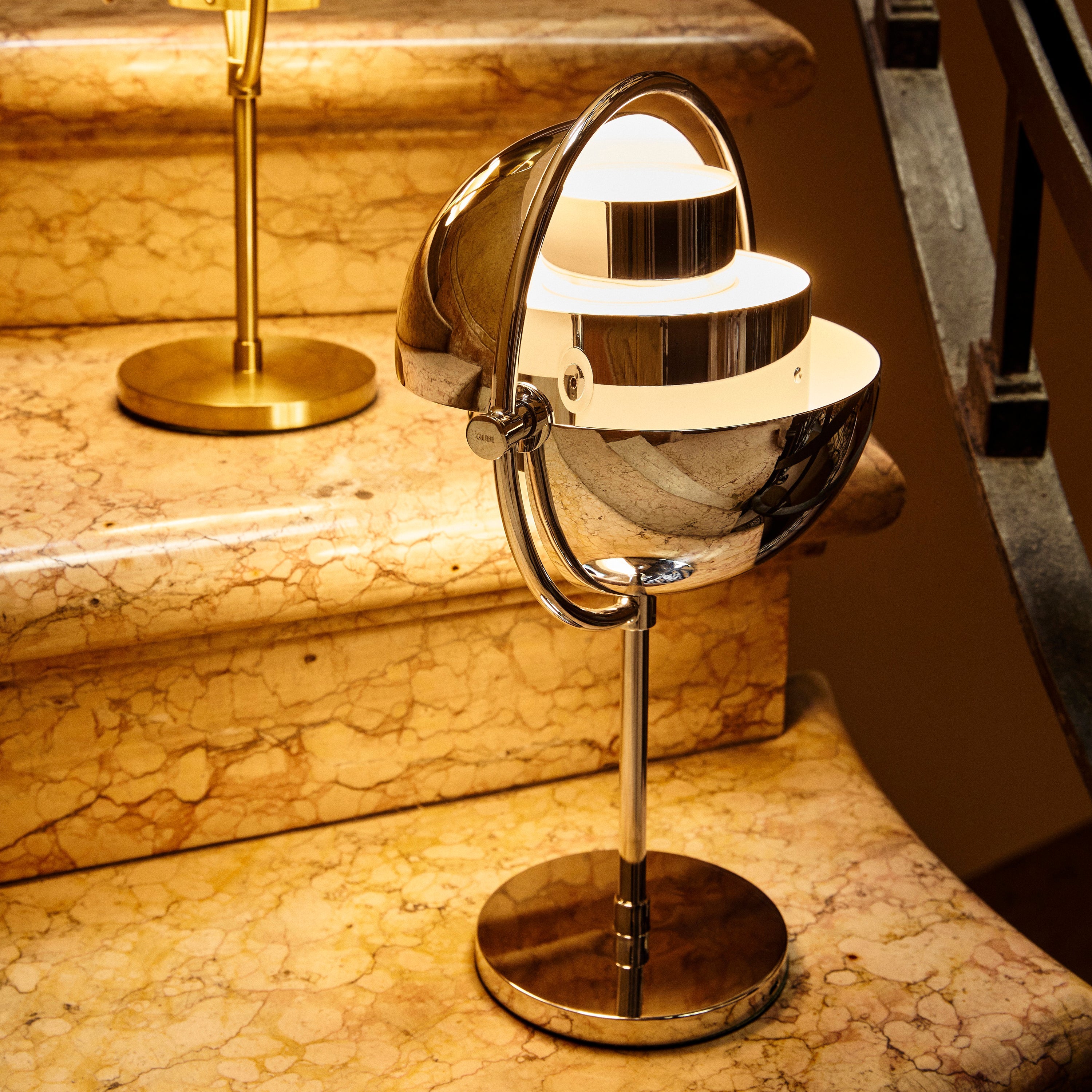 A glowing chrome table lamp with a rounded, tilting shade on a round base, sitting on marble stairs