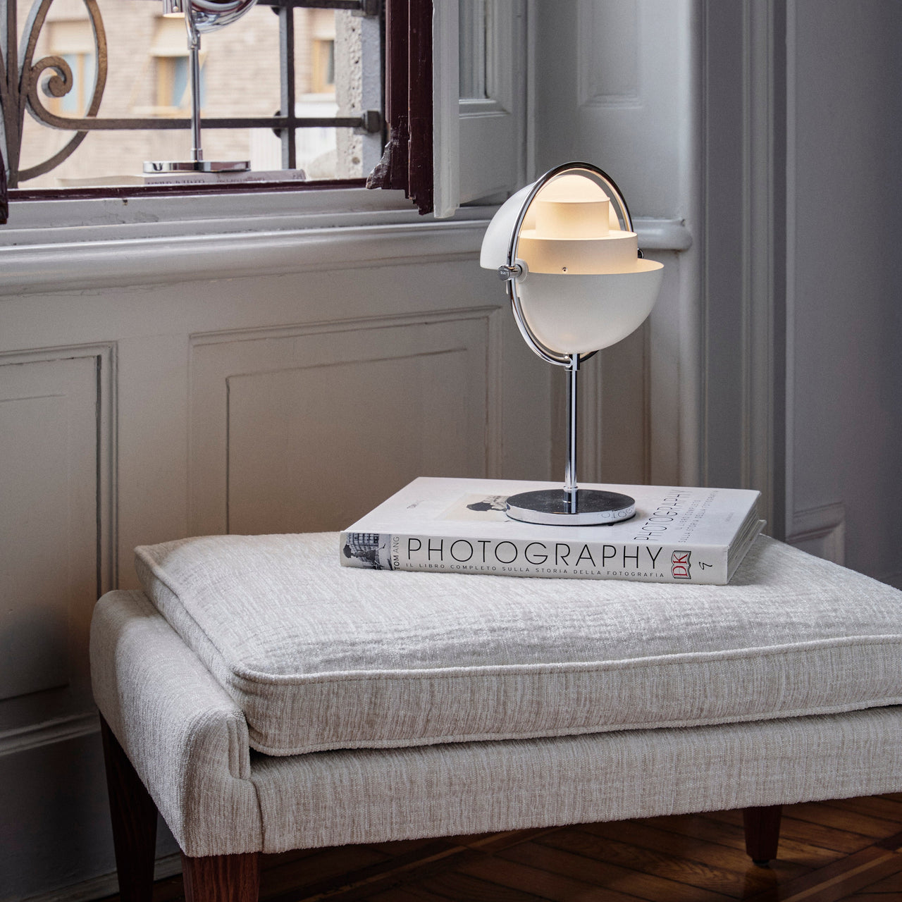 A white half-globe table lamp on a chrome stem and round base, sitting on a book on a cream upholstered bench by a window