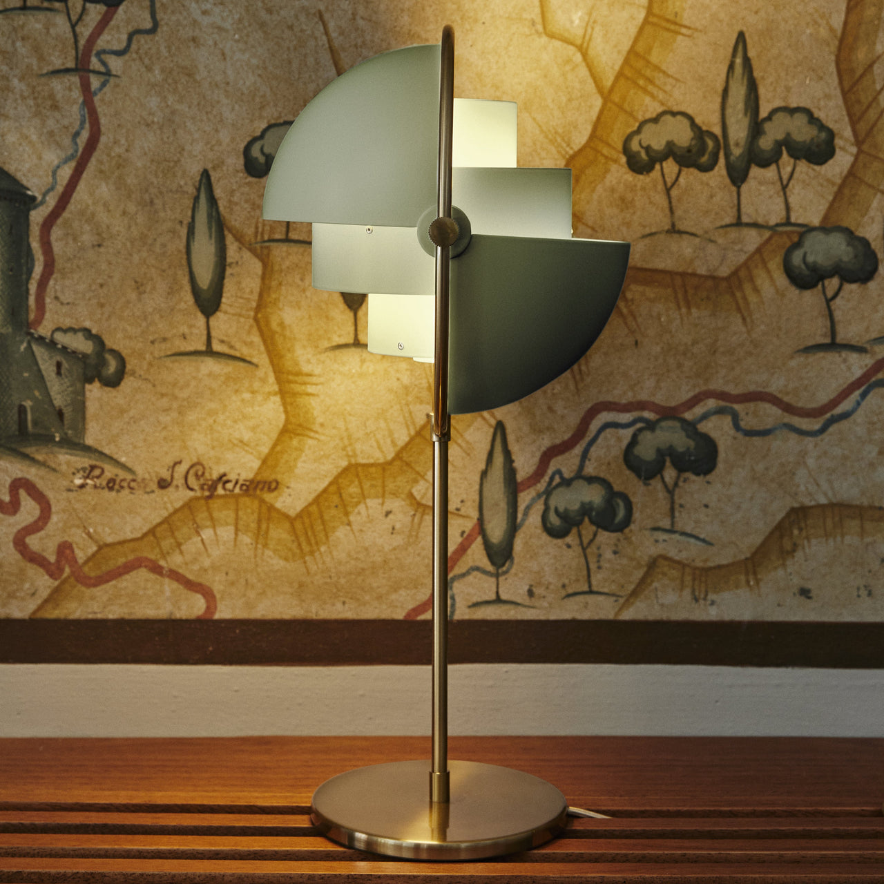 A table lamp with a brass base and stem and pivoting pale green quarter-circle shades, in front of a painted map mural