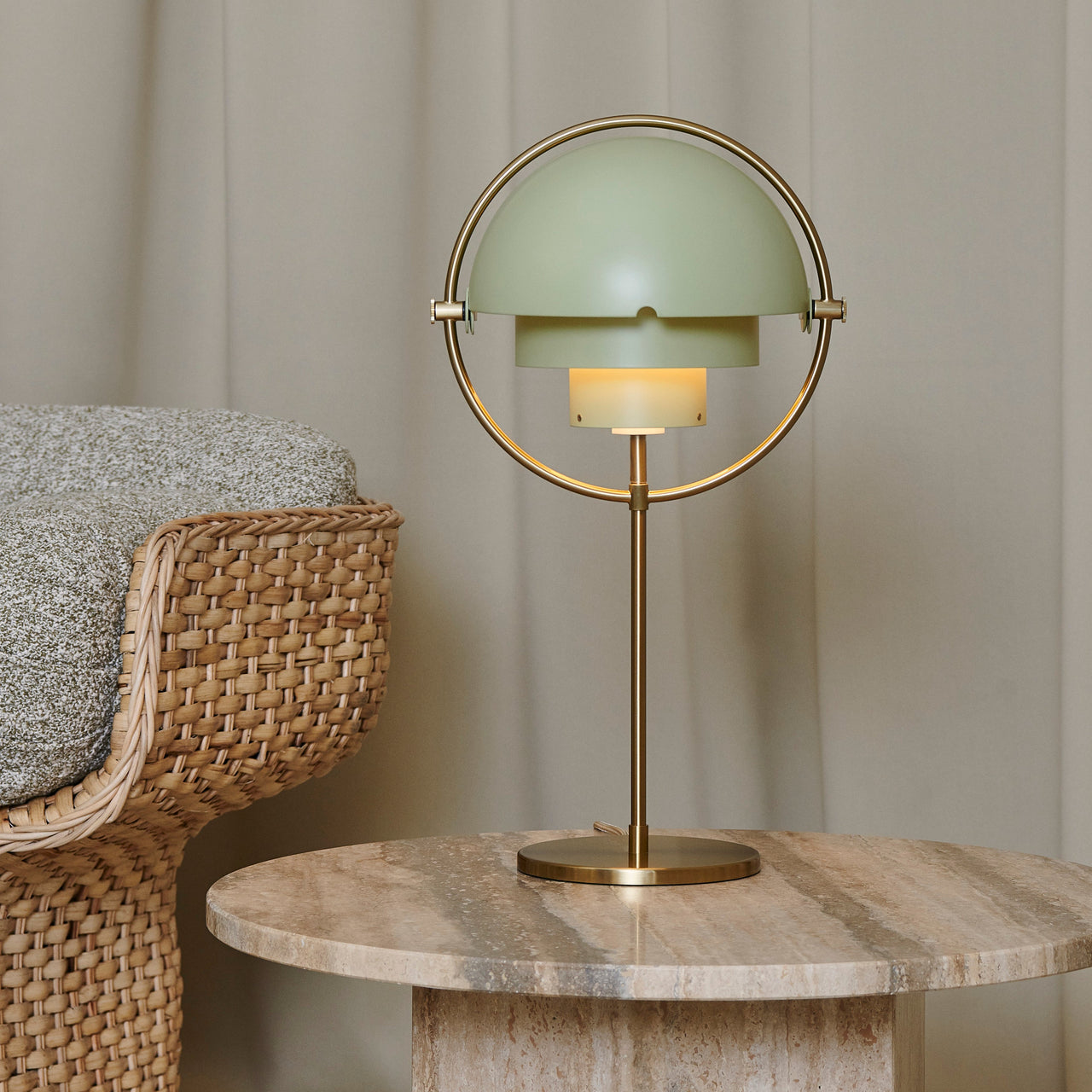 A table lamp with a pale green half-dome shade in a brass ring, on a stone side table by a woven chair
