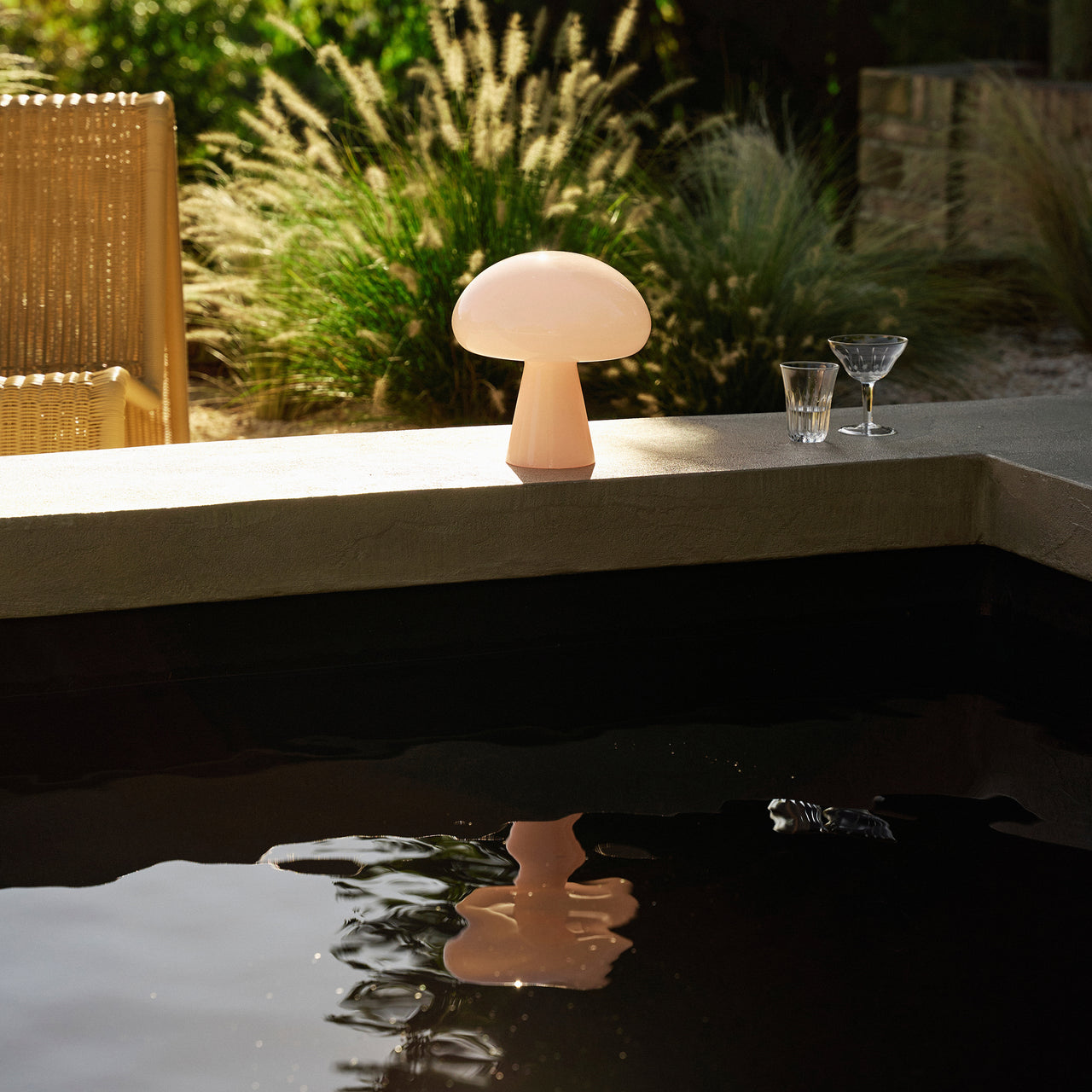A pale pink mushroom-shaped lamp glowing on a stone ledge beside a dark pool in a garden