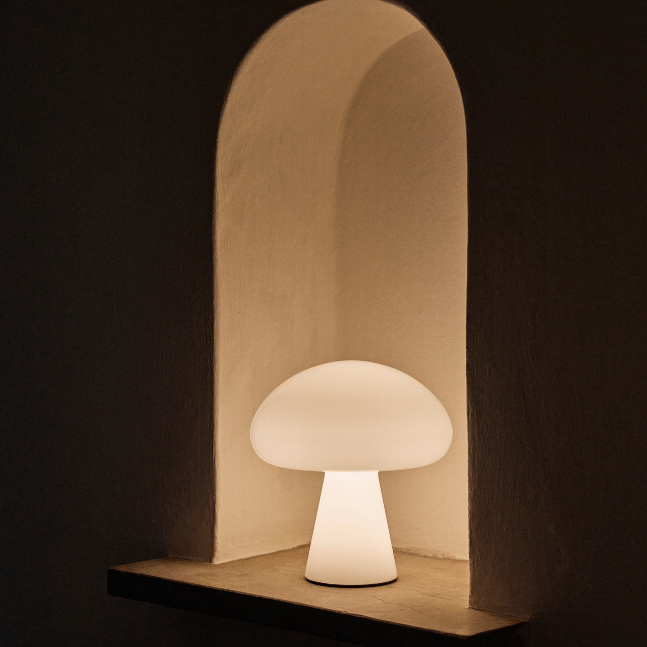 A glowing white mushroom-shaped lamp sits in an arched plaster wall niche in a dark room