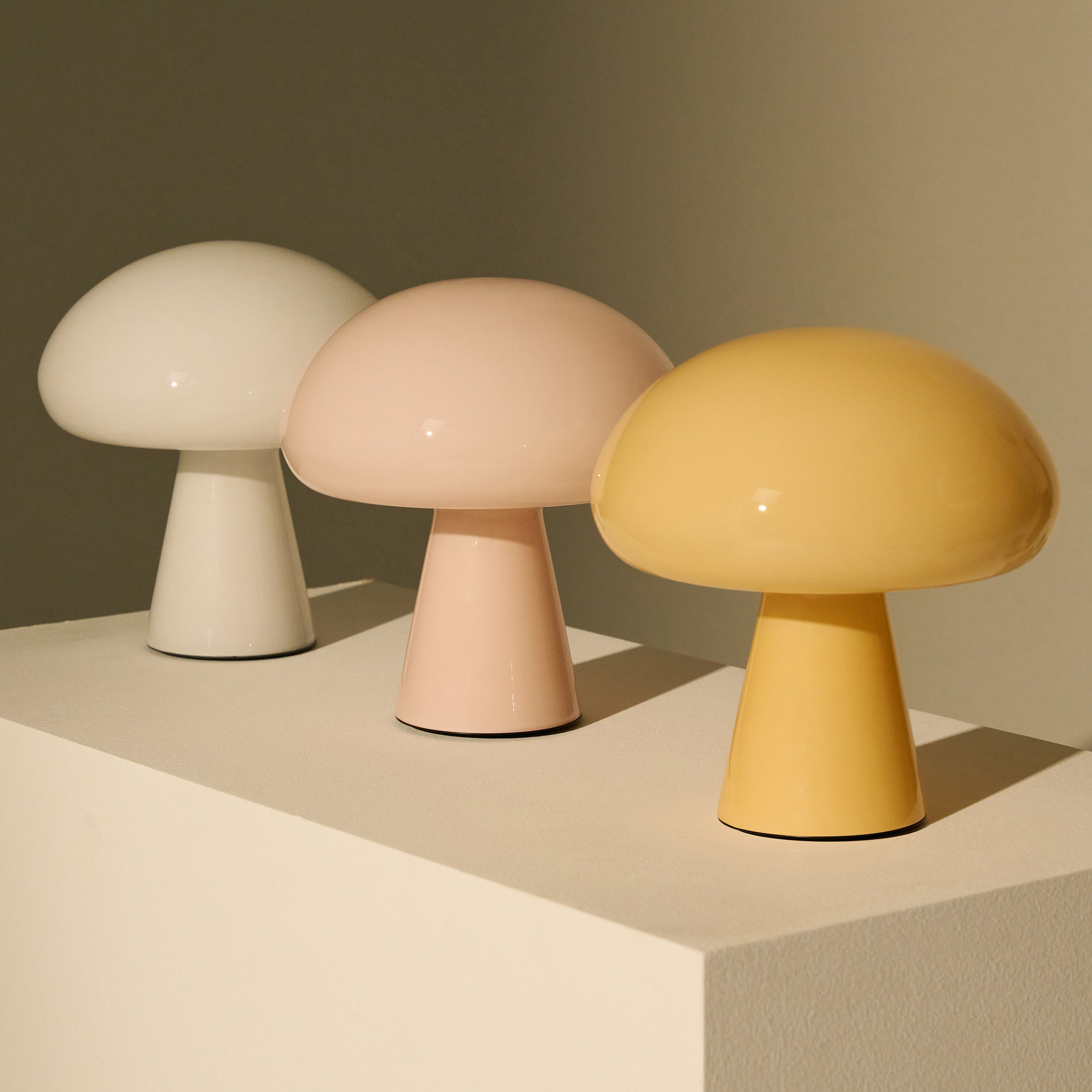 Three glossy mushroom-shaped table lamps in white, pale pink and yellow on a cream plinth