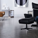 A black leather footstool on a four-star swivel base beside a matching wingback chair on a concrete floor