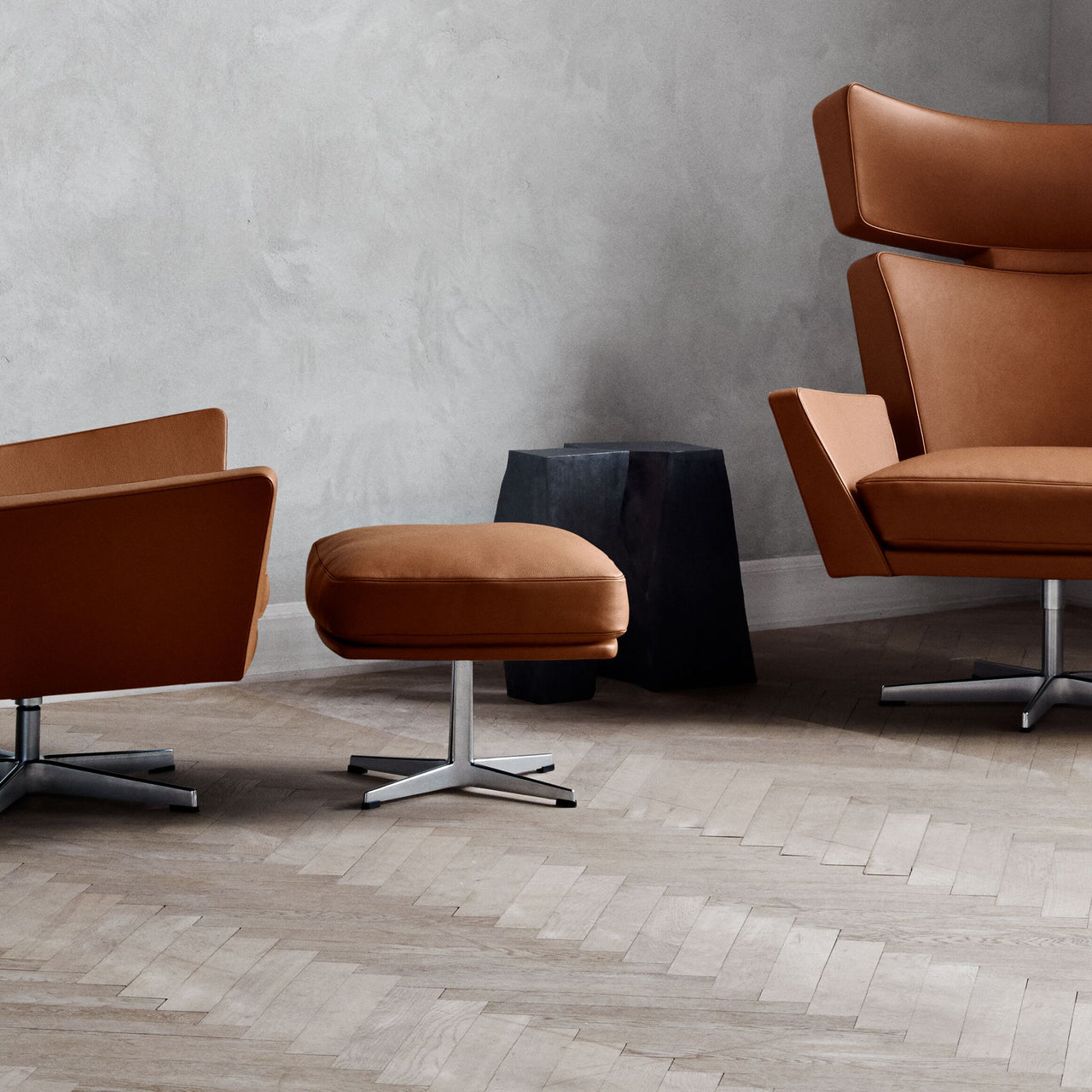 A tan leather footstool on a polished metal base beside two matching armchairs on a herringbone floor