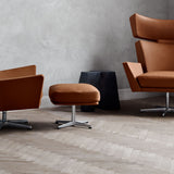 A tan leather footstool on a polished metal base beside two matching armchairs on a herringbone floor