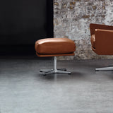 A tan leather footstool on a four-star polished metal base beside a matching armchair, against a brick wall