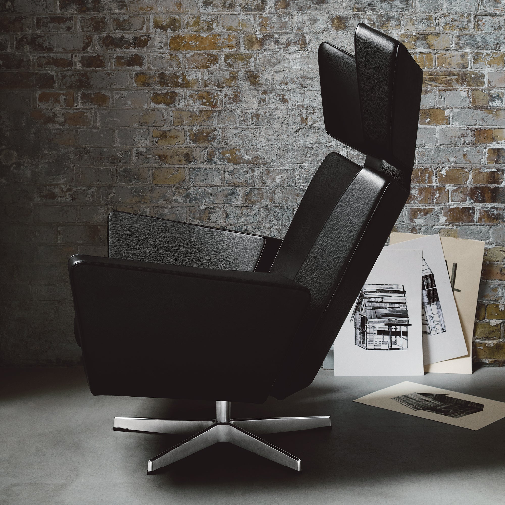 A black leather lounge chair with a tall winged headrest on a polished five-star swivel base, against a brick wall