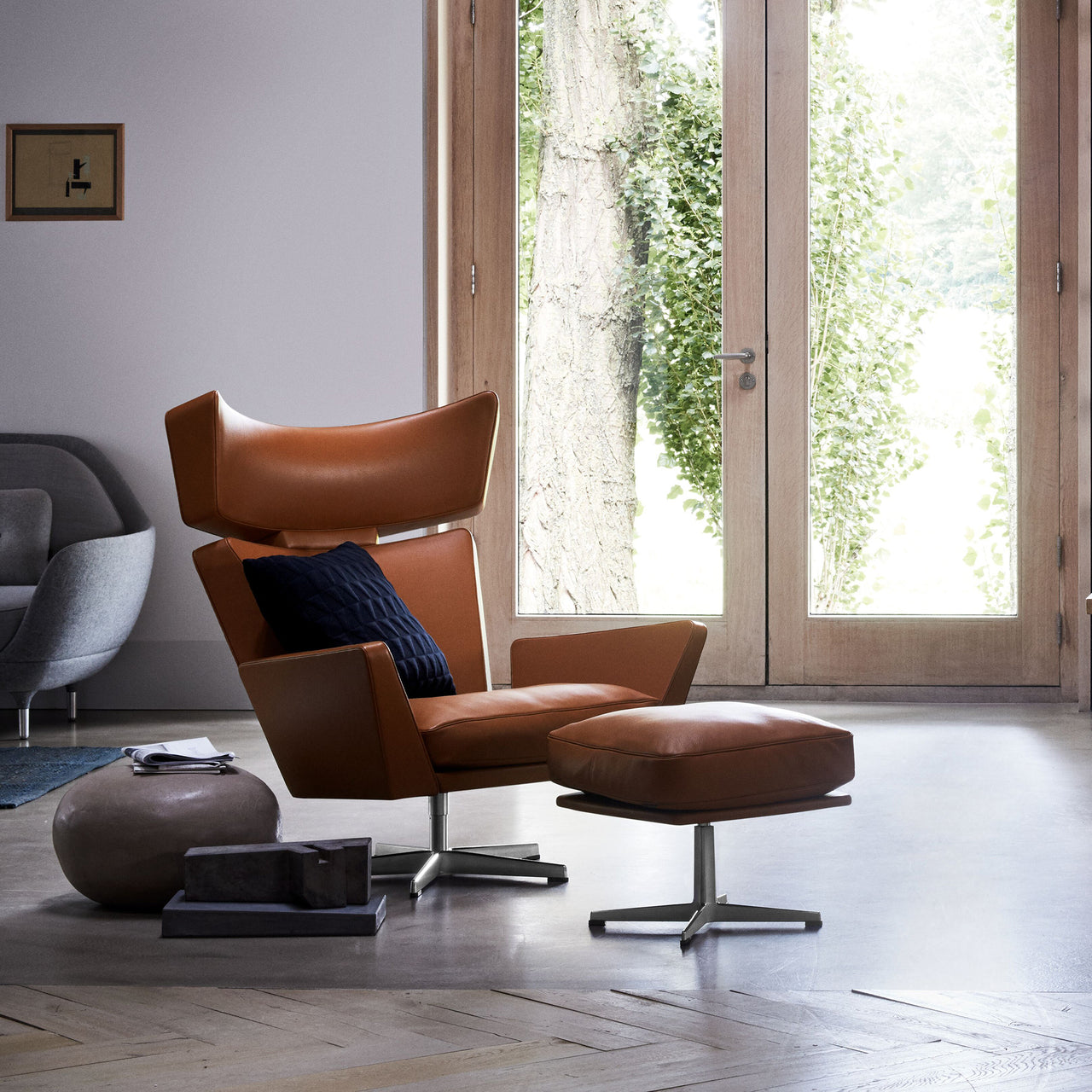 A tan leather wingback lounge chair with matching ottoman and metal swivel bases beside tall glass doors