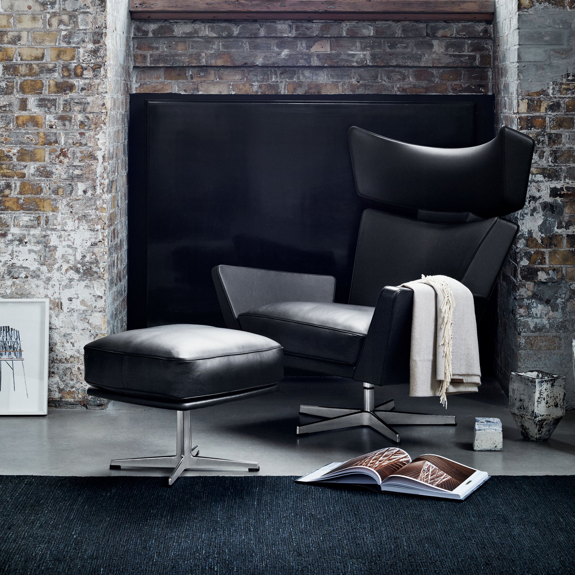 A black leather wingback lounge chair with matching ottoman on steel swivel bases, beside a brick wall