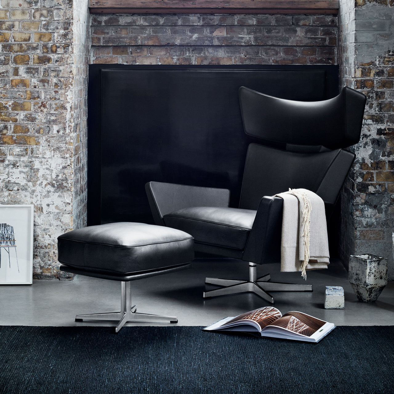 A black leather wingback lounge chair with matching ottoman on steel swivel bases, beside a brick wall