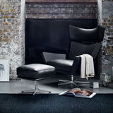 A black leather wingback lounge chair with matching ottoman on steel swivel bases, beside a brick wall