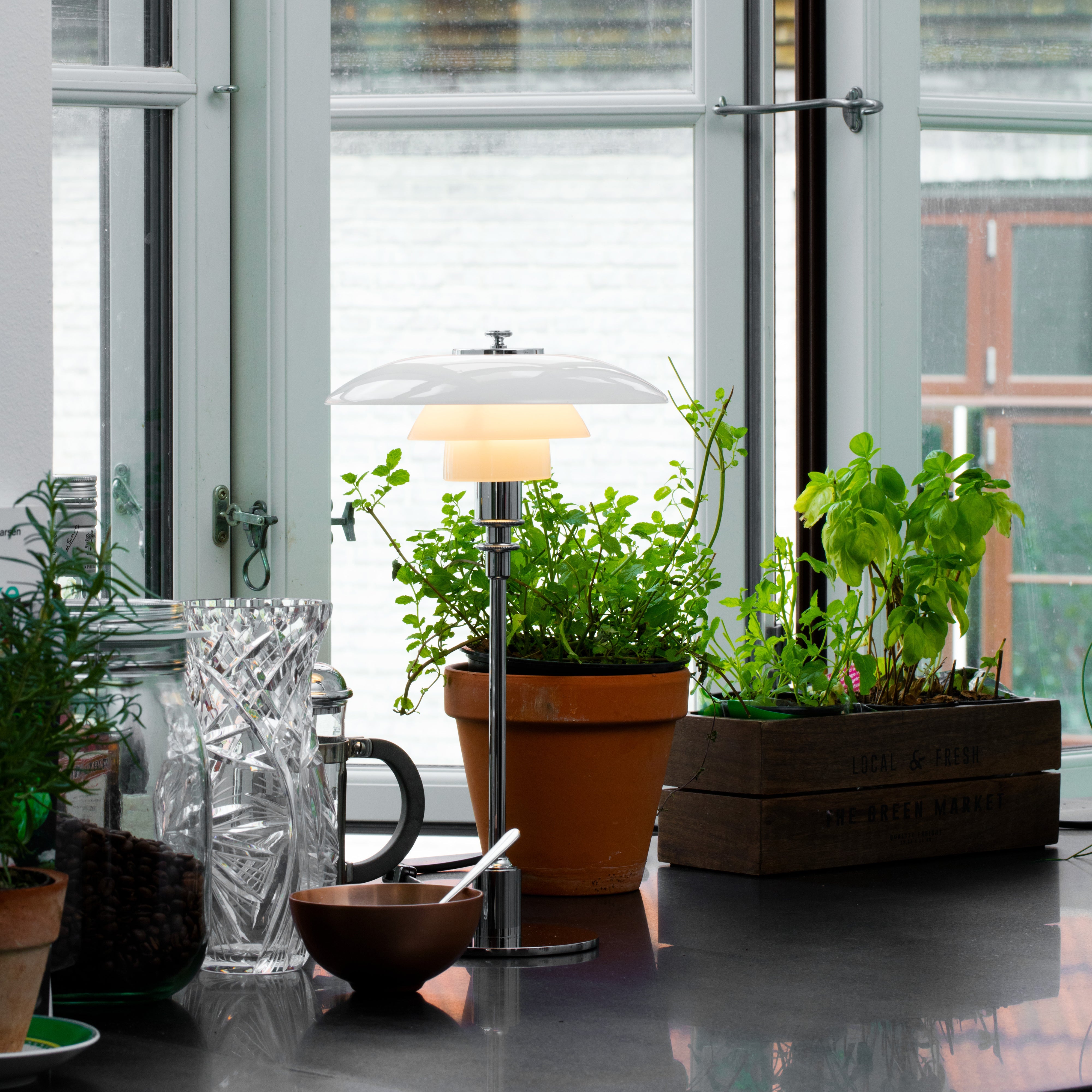 A glowing table lamp with layered white glass shades on a chrome stem, beside potted herbs by a window